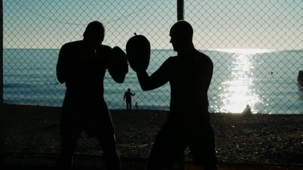 Siluetas. Dos atletas boxeadores masculinos están entrenando en un gimnasio al aire libre en una playa cerca del mar . — Vídeo de stock