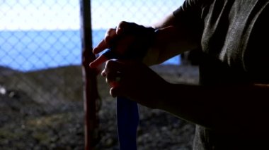 Yakın plan. Süper yavaş çekim. Silüet. Boksör maçtan önce elini elastik bir bandajla bandajlıyor. Sahildeki spor salonunda, mavi denizin üstünde.