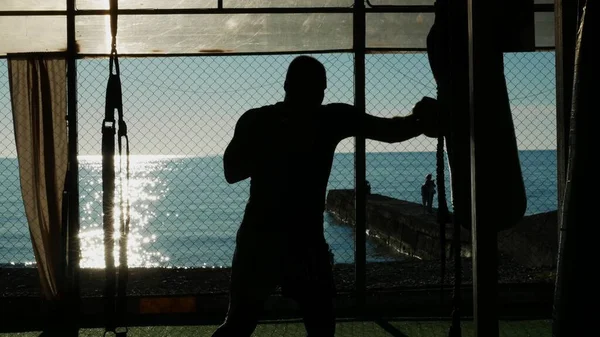 Silhueta. Boxer bate saco de perfuração em ginásio aberto na praia com vista para o mar . — Fotografia de Stock