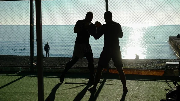 Silhuetter. Två manliga boxare idrottare tränar i en utomhus gym på en strand nära havet. — Stockfoto