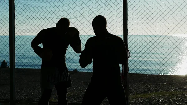 silhouettes. Two male boxer athlete are training in an outdoor gym on a beach near the sea.