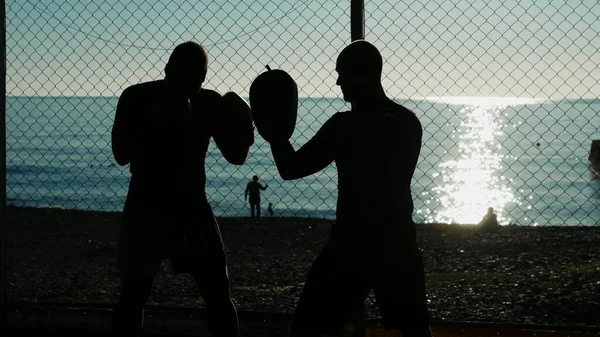 Sziluettek. Két férfi bokszoló atléta edz egy szabadtéri tornateremben egy tengerparti strandon.. — Stock Fotó