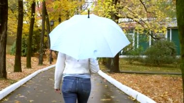 Beyaz deri ceketli, kot pantolonlu, ince bir kadın yağmurda mavi şemsiyeyle sonbahar parkında yürüyor.