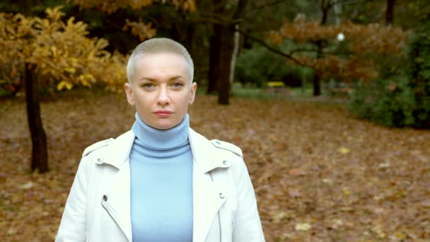 Κομψή ξανθιά γυναίκα με πολύ κοντά μαλλιά σε ένα λευκό δερμάτινο μπουφάν, το περπάτημα στο πάρκο φθινόπωρο. Κοιτάζοντας την κάμερα — Αρχείο Βίντεο