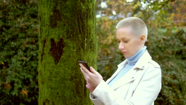 Una mujer de pelo corto utiliza un teléfono celular mientras está de pie junto a un árbol, en cuya corteza es visible un corazón — Vídeos de Stock