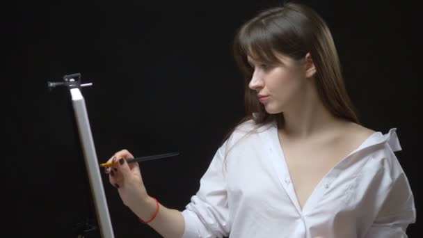 Cute girl artist paints a picture on canvas with oil paints. black background — ストック動画