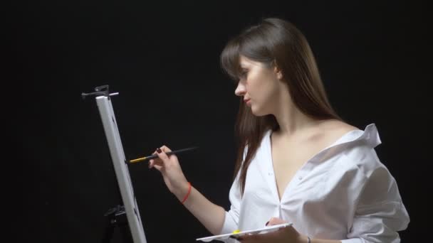 Cute girl artist paints a picture on canvas with oil paints. black background — Stock Video