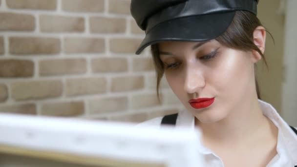 Girl artist in leather cap draws a picture on the canvas against a brick wall — Stock Video