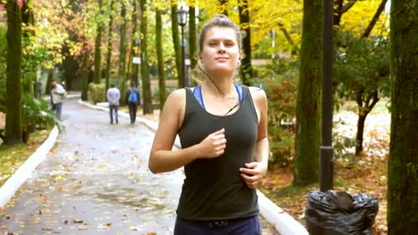 Sportliches Mädchen mit Kopfhörern joggt im Herbstpark — Stockvideo