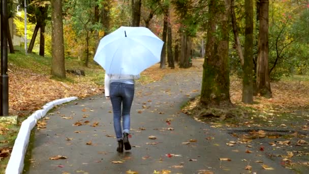 Fehér bőrdzsekis nő sétál az őszi parkban egy kék esernyővel — Stock videók