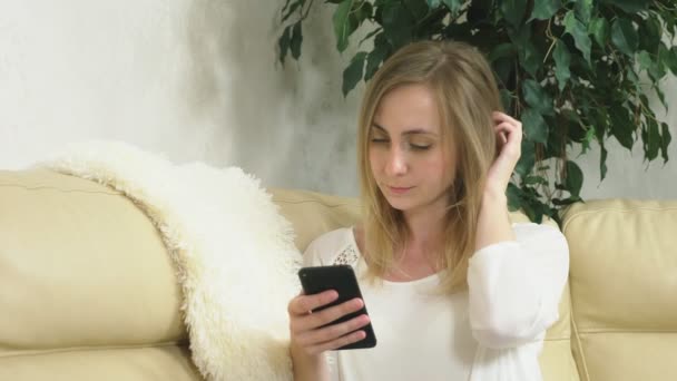 Διαταραγμένη κοπέλα χρησιμοποιεί ένα smartphone στο σπίτι κάθεται στον καναπέ. Άσχημα νέα. — Αρχείο Βίντεο