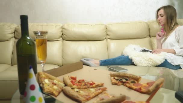 Sevimli sarışın kız kanepede kitap okuyor, şarap içiyor ve pizza yiyor. — Stok video