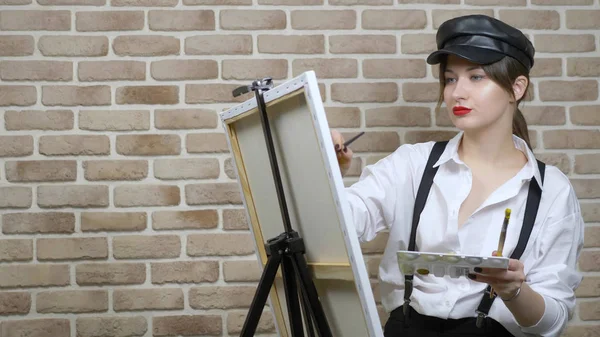 Girl artist in leather cap draws a picture on the canvas against a brick wall — Stock Photo, Image