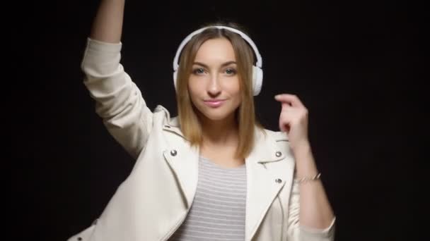 Linda chica en auriculares blancos y una chaqueta de cuero blanco sobre fondo negro — Vídeos de Stock