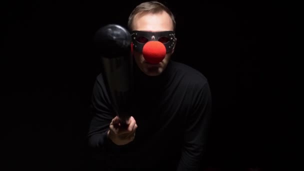 Homem em uma máscara de couro com um nariz de palhaço vermelho acenando um morcego em um fundo preto — Vídeo de Stock