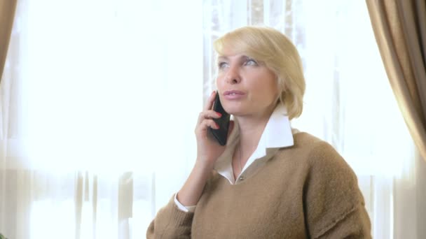 Красивая женщина разговаривает по мобильному телефону, стоит в офисе у окна — стоковое видео