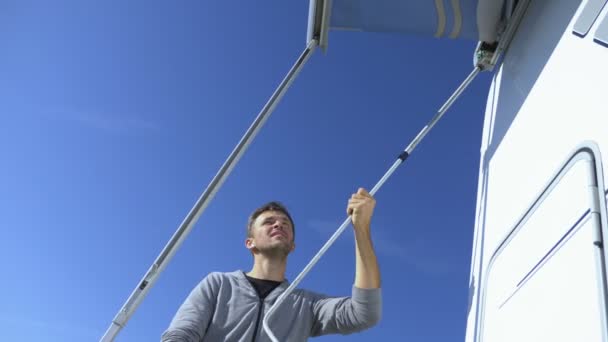 Un hombre coloca un dosel del sol en una casa rodante contra un cielo azul claro — Vídeo de stock
