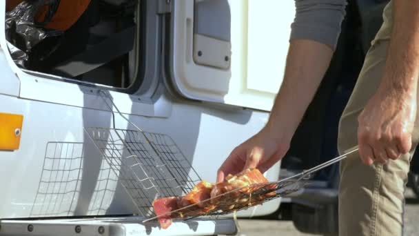 Чоловік готує м'ясо на грилі на відкритому повітрі на фоні моторів — стокове відео