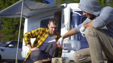 İki adam, doğada karavanın yakınındaki ızgarada et pişiriyor.