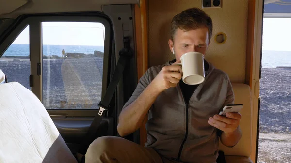 Człowiek pije kawę i używa smartfona siedzącego w przyczepie kempingowej nad morzem — Zdjęcie stockowe