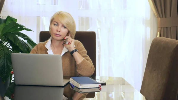 Жінка в офісі говорить по телефону, дивлячись на ноутбук — стокове фото