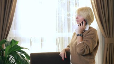 Güzel bir kadın cep telefonuyla konuşuyor, ofiste pencerenin yanında duruyor.