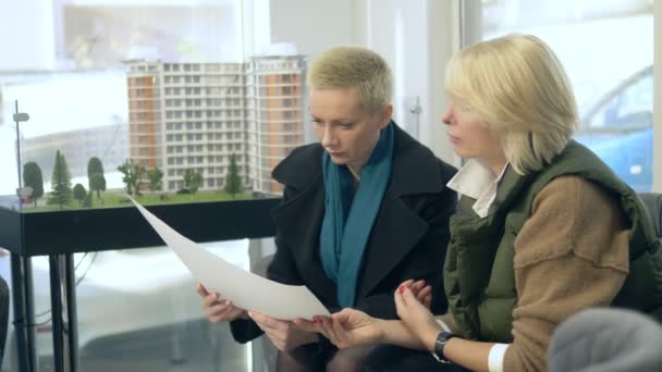 Две женщины разговаривают, сидя в офисе продаж на фоне планировки здания — стоковое видео