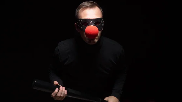 Человек в кожаной маске с красным клоунским носом машет битой на черном фоне — стоковое фото