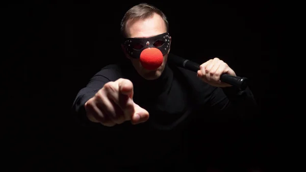 Homem em uma máscara de couro com um nariz de palhaço vermelho acenando um morcego em um fundo preto — Fotografia de Stock
