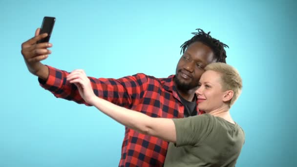 Afrikaanse jongen en Kaukasisch meisje maken selfie met smartphone. blauwe achtergrond. — Stockvideo