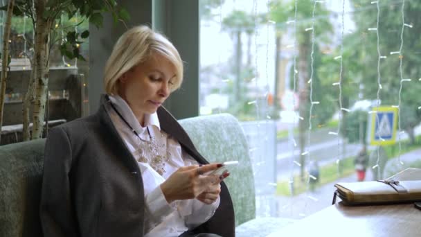 Elegante mujer rubia con estilo utilizando el teléfono móvil sentado en un café — Vídeos de Stock