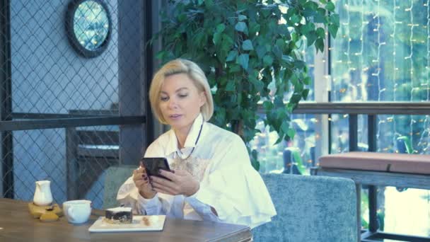 Κομψή κομψή ξανθιά γυναίκα που χρησιμοποιεί το κινητό τηλέφωνο κάθεται σε ένα καφέ — Αρχείο Βίντεο
