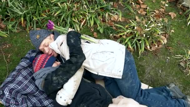 Άστεγος άντρας και γυναίκα ξαπλώνουν σε χαρτόκουτο σε ένα πάρκο κοντά σε κήπο με λουλούδια. — Αρχείο Βίντεο