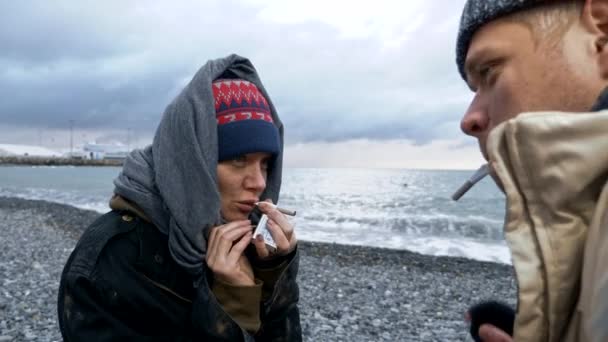 Pareja sin hogar, hombre y mujer fumando en la orilla del mar — Vídeo de stock