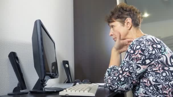 Elderly woman at the computer sits at a table in the home office — 비디오