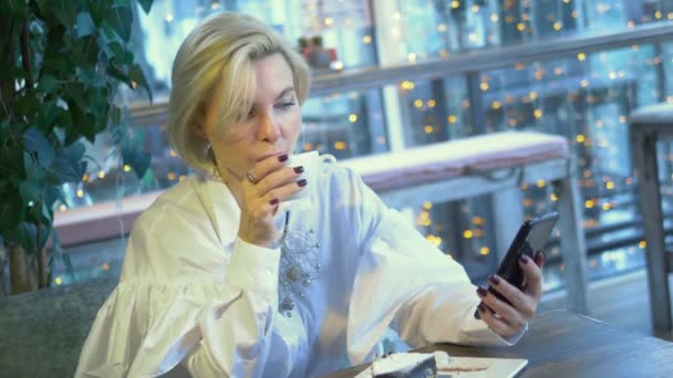 Elegante mujer rubia con estilo utilizando el teléfono móvil sentado en un café — Vídeo de stock