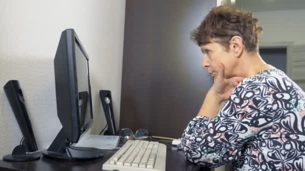 Elderly woman at the computer sits at a table in the home office — 비디오