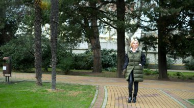 Sonbaharda bir şehir parkında yürüyen şık, şık, sarışın bir kadın.