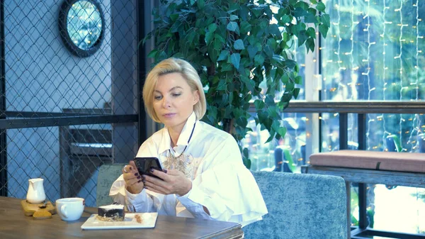 Wanita pirang elegan bergaya menggunakan ponsel duduk di kafe — Stok Foto