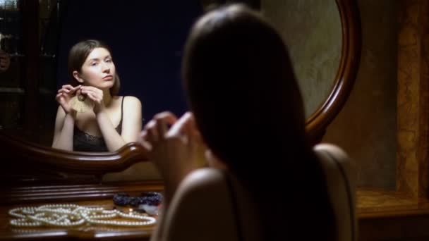 Schöne Mädchen zieht Ohrringe an, sitzt in der Abenddämmerung vor dem Spiegel. — Stockvideo
