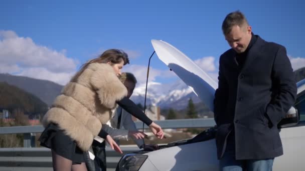 Két nő javítja a kocsit. Egy férfi áll a közelben és nézi őket. — Stock videók