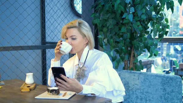 Elegante stijlvolle blonde vrouw met behulp van mobiele telefoon zitten in een cafe — Stockfoto