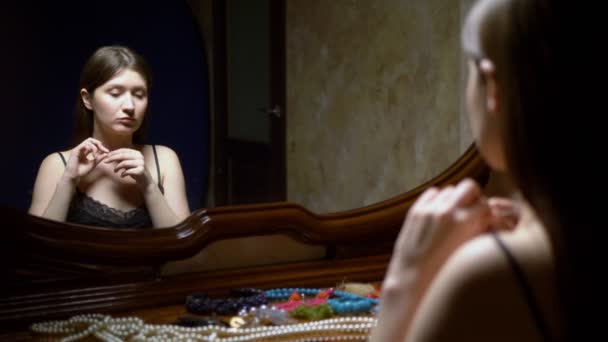 Beautiful girl puts on earrings, sitting in front of the mirror at dusk. — Stock Video