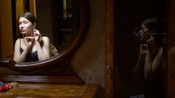 Красивая девушка надевает серьги, сидя перед зеркалом в сумерках . — стоковое видео