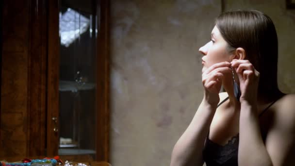 Beautiful girl puts on earrings, sitting in front of the mirror at dusk. — Stock Video