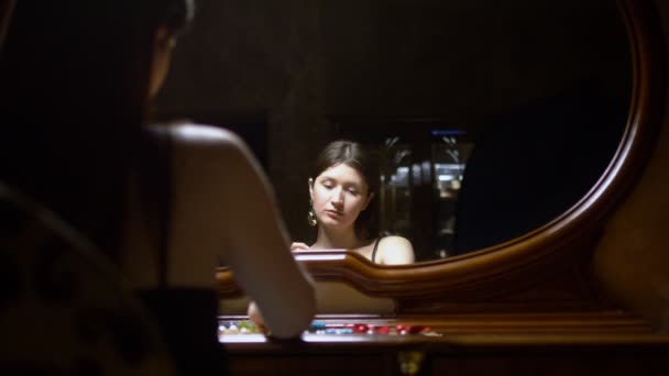 Красива дівчина одягає сережки, сидячи перед дзеркалом в сутінках . — стокове відео