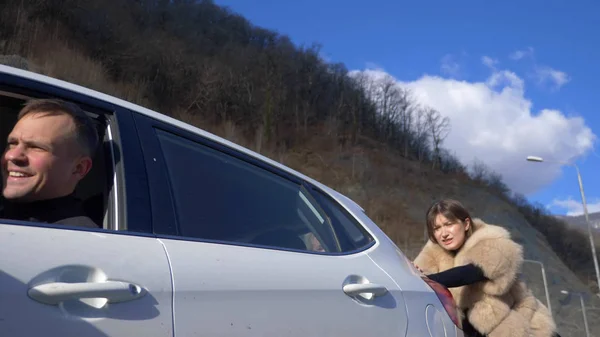 Duas meninas bonitas empurrar o carro quebrado na estrada. um homem senta-se ao volante — Fotografia de Stock