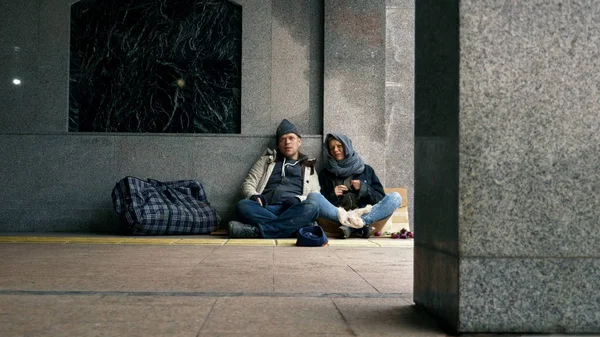 Бездомные просят милостыню, сидя на тротуаре у стены. — стоковое фото