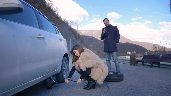 Umorismo. donna che cambia una ruota dell'auto. uomo che parla al telefono — Foto Stock