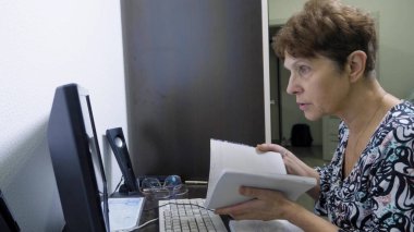 Bilgisayarın başındaki yaşlı kadın ofisteki bir masada oturuyor.